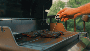 Meat thermometer checking cooked ribs on a grill for perfect barbecue temperature.