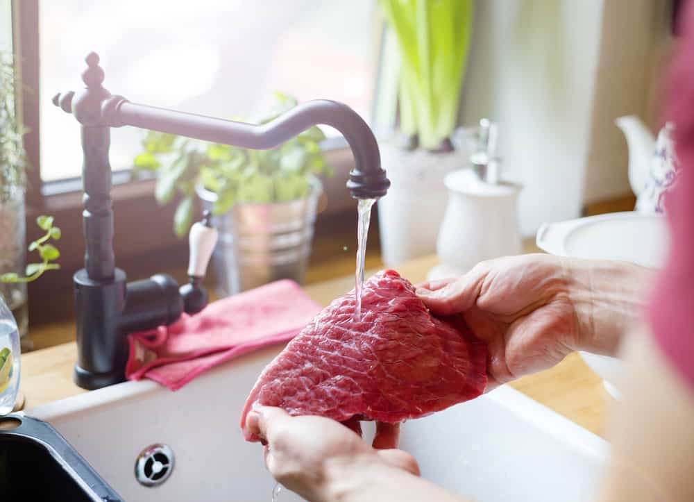 Fresh beef being washed under running water in a kitchen sink for food safety and hygiene.