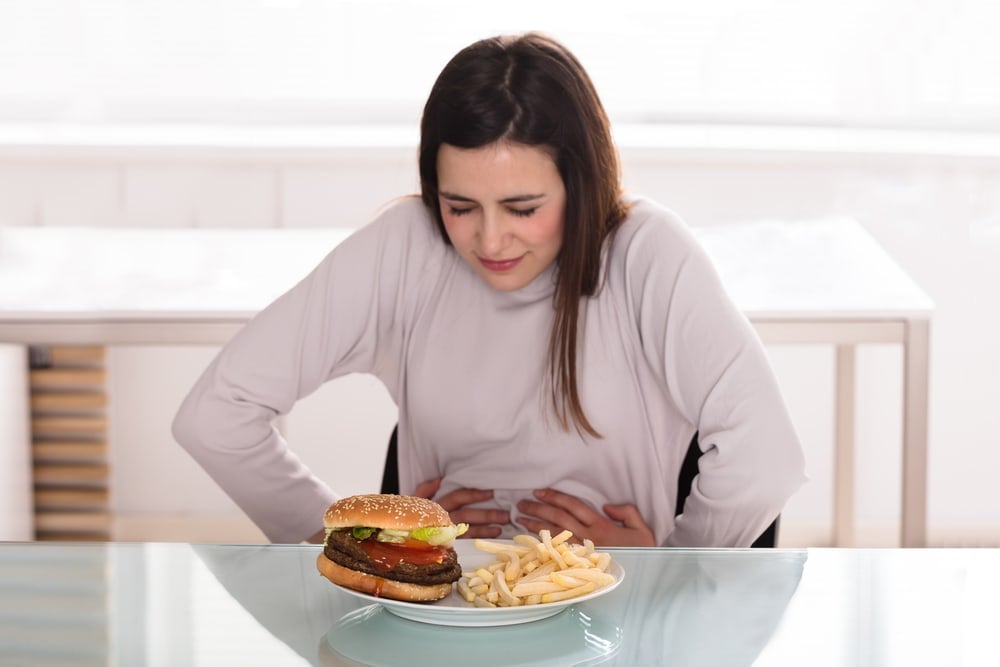 Nauseated woman with stomach ache looking at burger and fries, food discomfort, food poisoning, digestive issues.