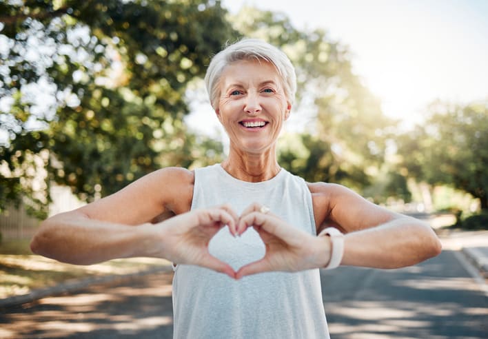 Caring elderly woman making heart shape outdoors, promoting health and wellness, autumn park background.