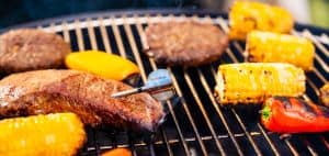 Juicy steaks grilling with corn, peppers, and a meat thermometer on a barbecue grill.